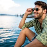 Brunette man wearing a green shirt and sunglasses smiles by a lake with healthier gums in Denver, CO