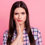 brunette woman with sensitive teeth cringes in pain and touches her cheek against a pink background
