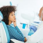 Black woman touches her face and talks to her dentist while sitting in a dental chair